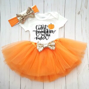 May include: A white baby bodysuit with the text "Cutest pumpkin in the patch" and an orange pumpkin graphic. The outfit includes an orange tutu skirt and a matching headband, both with gold sequin bows. The outfit is designed for babies.