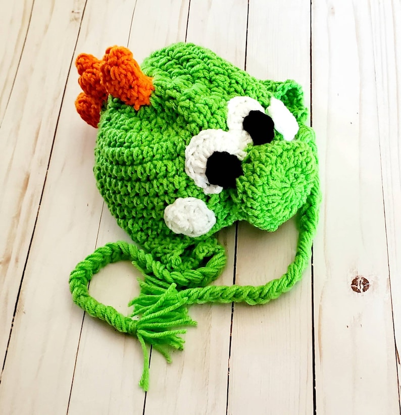 May include: A bright green crocheted hat featuring a character design with large white and black eyes, a white snout, and a row of orange spikes along the top. The hat includes two long, braided green yarn ties with fringed ends at the bottom.