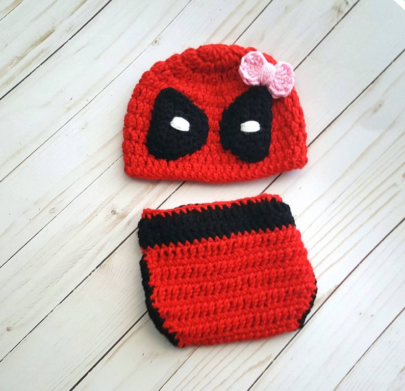 May include: Red and black crocheted baby hat and diaper cover with a black eye mask and a pink bow. The hat and diaper cover are made to look like the superhero Deadpool.