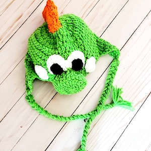 May include: A bright green crocheted hat featuring a creature design with a prominent orange dorsal fin, large white and black eyes, and two braided green earflaps with tassels. The hat is constructed with a textured yarn stitch pattern.