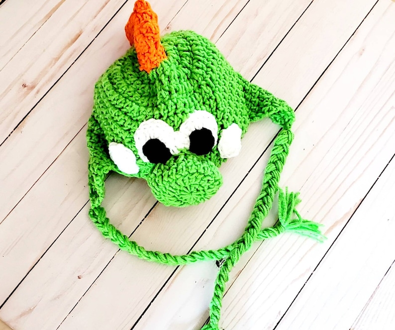 May include: A bright green crocheted hat featuring a creature design with a prominent orange dorsal fin, large white and black eyes, and two braided green earflaps with tassels. The hat is constructed with a textured yarn stitch pattern.