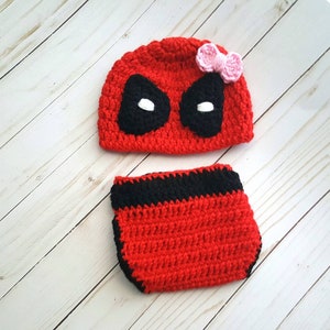 May include: A red and black crochet baby set, including a beanie and diaper cover. The beanie features black eye shapes and white eye accents, with a pink bow. The diaper cover has a black band.