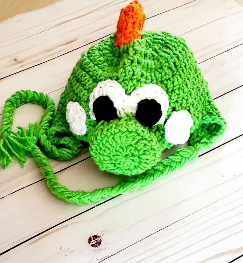 May include: A bright green crocheted hat featuring a character design with a prominent orange spike on top, large white eyes with black pupils, and two braided green ties hanging from the sides. The hat has a textured, yarn-based construction.