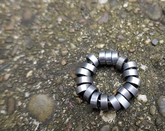 Puede incluir: Un objeto circular de metal compuesto por pequeños cilindros plateados dispuestos en forma de anillo. El objeto descansa sobre una superficie de piedra texturizada gris y marrón. Los cilindros parecen ser de metal brillante.