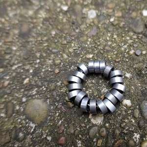 May include: A circular metal object composed of small, silver-colored cylinders arranged in a ring. The object is resting on a textured, gray and brown stone surface. The cylinders appear to be made of a shiny metal.