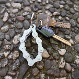 Op de afbeelding: Een zilveren fietsketting sleutelhanger met een zwarte sleutel met de tekst "ONGUARD" en een gouden sleutel met de tekst "BRICARD".