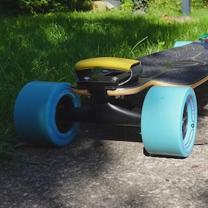 May include: A longboard skateboard with a black grip tape deck, light blue wheels, and a yellow handle. The wheels are a vibrant blue, and the board is set against a background of green grass and concrete.