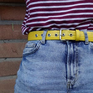 Könnte beinhalten: Nahaufnahme eines gelben Gürtels mit silberner Schnalle und Ösen, getragen mit blauen Jeansshorts. Der Gürtel wird mit einem bordeauxrot-weiß gestreiften Hemd kombiniert. Der Gürtel ist etwa 3,8 cm breit.