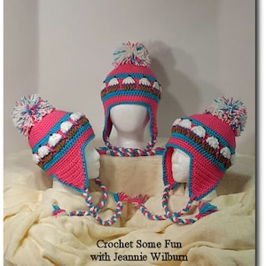 May include: Three pink crocheted hats with white and blue accents. Each hat has a white pom-pom on top and a long white and blue crocheted cord. The hats are arranged on a white fluffy surface. The text "Crochet Some Fun with Jeannie Wilburn" is at the bottom of the image.