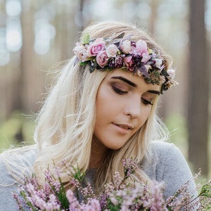 May include: A woman with long blonde hair wears a flower crown with pink and purple flowers. She is holding a bouquet of purple flowers.
