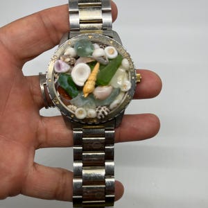 May include: A silver and gold wristwatch with a unique face. The face is filled with small seashells and sea glass.