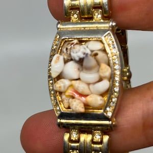 May include: A gold and silver tone watch with a rectangular face. The face is filled with small seashells.