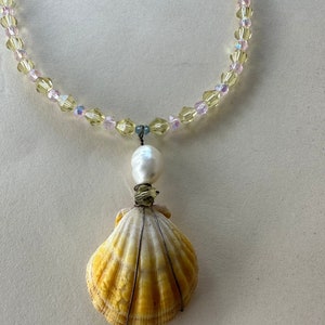 Op de afbeelding: Een gele schelp ketting met een parel en kristallen kralen. De schelp is omwikkeld met draad en de ketting is gemaakt van heldere en roze kristallen kralen.