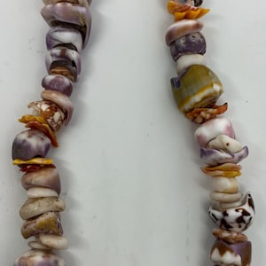 May include: A necklace composed of assorted seashells in hues of purple, white, yellow, and brown. The shells are threaded together, forming a unique piece of natural jewellery. The necklace is shown against a white backdrop.