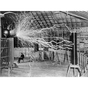 Könnte beinhalten: Ein Schwarzweißfoto einer großen elektrischen Spule in einem Holzgebäude. Die Spule erzeugt einen großen elektrischen Lichtbogen, der zur Decke reicht. Ein Mann sitzt im Vordergrund auf einem Stuhl und beobachtet den Lichtbogen.