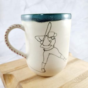 May include: Cream-colored ceramic mug with a baseball theme. The mug features a black line drawing of a baseball player in a batting stance. The handle is designed to resemble a baseball with red stitching. The rim is teal.