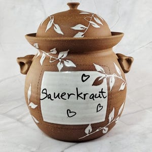 Può includere: Vaso per crauti in ceramica marrone con coperchio, design a foglie bianche e la scritta "Sauerkraut" su un'etichetta bianca. Il vaso ha due manici e cuori.