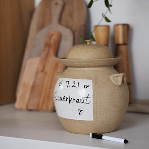 May include: A beige ceramic jar with a lid and a white label that says "7.21 Sauerkraut" in black marker. The jar is sitting on a white surface with a white marker next to it.