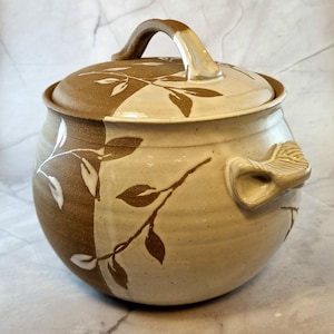 May include: A handcrafted ceramic jar with a lid, featuring a brown and cream color scheme. The jar is adorned with leaf and branch designs. It has a handle on the lid and two side handles. The jar is approximately 8 inches in height and 8 inches in diameter.
