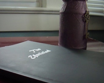 May include: A black rectangular book with the white text The Untitled printed on the cover, positioned next to a purple textured jar containing dark feathers and adorned with a hanging antique brass-colored skeleton key ornament.