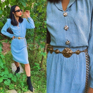 May include: A light blue dress with a textured fabric and a gold chain belt with large circular medallions. The dress has a collared neckline and long sleeves. The dress is worn by a person with long black hair and sunglasses.
