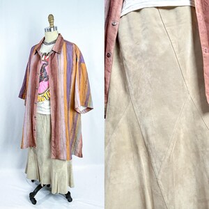 May include: A mannequin showcases a layered ensemble. A long, button-down shirt with vertical stripes in orange, purple, and brown is worn over a white graphic tee and a beige suede skirt. A pink jacket is draped over the shoulders.