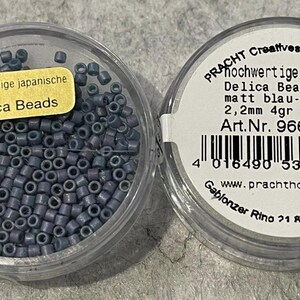 Könnte beinhalten: Zwei transparente Kunststoffbehälter gefüllt mit matten blau-grauen Delica-Perlen. Ein Behälter hat ein gelbes Etikett mit der Aufschrift "hochwertige japanische Delica Beads". Der andere hat ein Etikett mit deutschem Text.