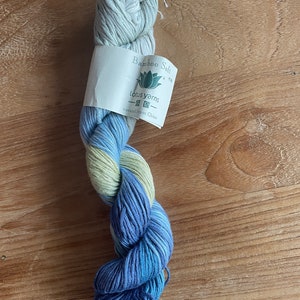 Könnte beinhalten: Ein Strang Bamboo Soft Garn in Blau-, Grün- und Weißtönen. Das Garn ist mit "Lotus Yarns" und "natural from China" gekennzeichnet. Das Garn ist gezwirnt und liegt auf einer Holzoberfläche.