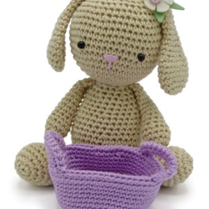 Könnte beinhalten: Ein gehäkeltes beigefarbenes Häschen mit rosa Nase und schwarzen Augen sitzt und hält einen lilafarbenen gehäkelten Korb.