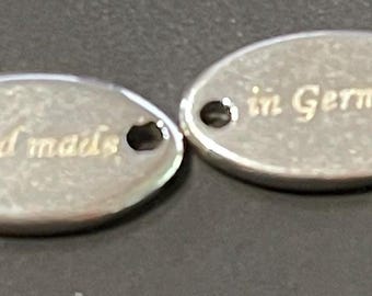 Könnte beinhalten: Zwei ovale Metallanhänger in Silber. Auf einem Anhänger steht "Hand made" in goldfarbenen Buchstaben. Auf dem anderen Anhänger steht "in Germany" in goldfarbenen Buchstaben. Jeder Anhänger hat ein kleines Loch oben.