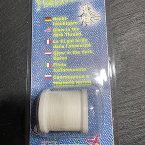 Könnte beinhalten: Eine weiße Spule mit Leuchtgarn zum Basteln. Die Verpackung zeigt eine Hexe und den Text "Halloween" in einer dekorativen Schrift. Der Text "Glow in the dark" ist in mehreren Sprachen geschrieben.