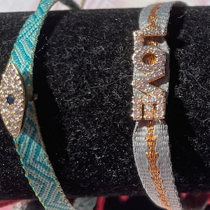 Könnte beinhalten: Zwei Armbänder mit gewebtem Design. Ein Armband ist türkis und weiß mit einem mit Diamanten besetzten bösen Augen-Anhänger. Das andere Armband ist grau und weiß mit einem mit Diamanten besetzten Anhänger, auf dem "LOVE" steht.