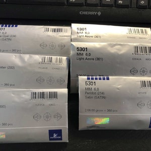 May include: Five packages of Swarovski crystals, each with a different color and name. The packages are labeled with the product number 5301, the size 6.0 mm, and the color and name of the crystal. The packages are all white with black text and a small Swarovski logo in the bottom right corner.