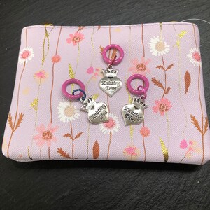 Könnte beinhalten: Drei violette und silberne Strickmarkierer mit einem herzförmigen Anhänger, auf dem "Knitting Diva" steht. Die Markierer befinden sich auf einer violetten und weißen Stofftasche mit Blumenmuster.