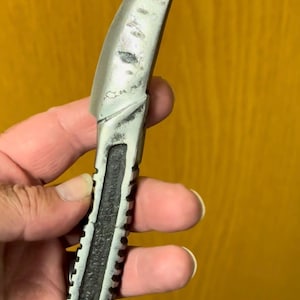 May include: A small, curved knife with a silver blade and a textured, black handle. The blade has a slight curve and a pointed tip. The handle has a series of ridges along its length. The knife is held against a yellow background.