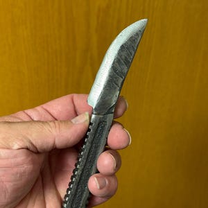 May include: A hand-forged, small knife with a curved blade and a textured, ridged handle. The blade is silver, and the handle is dark gray. The knife appears to be made of metal and is held in a person's hand.