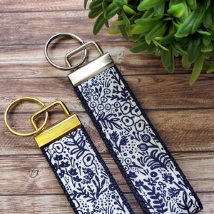 May include: Two keychains with a white background and a navy blue floral pattern. The keychains have gold metal rings and a navy blue trim.