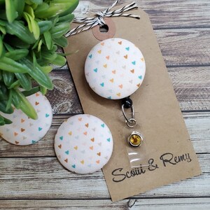 May include: A white fabric button with a pattern of small orange, teal, and white hearts. The button is attached to a retractable badge holder with a clear plastic case and a gold-colored clasp. The badge holder is attached to a brown card with the text "Scotti & Remy".