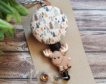 May include: A badge reel featuring a circular fabric button with a multi-colored forest tree pattern, attached to a brown silicone moose charm. The reel includes a clear plastic strap with a metal snap and a small amber rhinestone, mounted on a card.