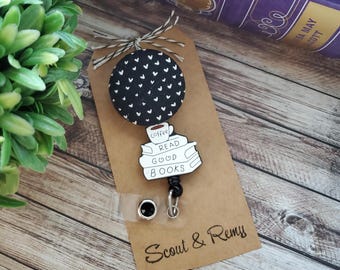 May include: A black and white badge reel with a black and white heart pattern. The badge reel has a white and black graphic of a coffee cup on top of a stack of books with the words "Read Good Books". The badge reel is attached to a brown tag with the words "Scout & Remy".