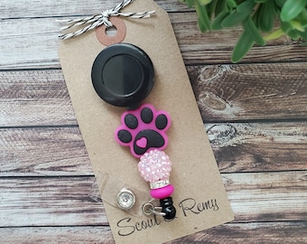 Può includere: Un portabadge retrattile nero e argento con un ciondolo di cristallo trasparente, un ciondolo a forma di zampa rosa con un ciondolo a forma di cuore rosa e un'etichetta marrone con il testo "Scout & Remy".