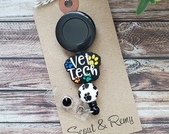 Può includere: Un porta badge retrattile nero con un ciondolo a forma di impronta di zampa bianco e nero. Il ciondolo ha le parole "Vet Tech" su di esso. Il porta badge è attaccato a una carta marrone con il testo "Scout & Remy".