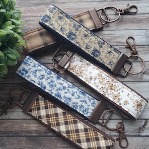 May include: Several fabric key fobs with different patterns, including floral and plaid designs. Each fob has a metal clasp and ring for keys. The colour scheme features blues, browns, and white, with a rustic aesthetic.