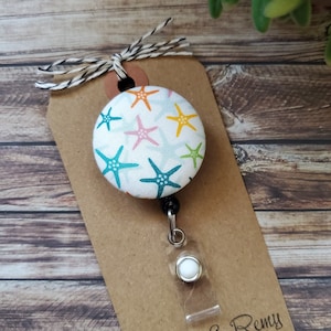 May include: A white button with a blue background and colorful starfish pattern attached to a black retractable badge holder with a white plastic clip. The button is attached to a brown card with the text "Scout & Remy" printed on it.
