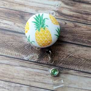 May include: A white fabric badge reel with a pineapple pattern. The reel has a green gem on the end of the retractable cord.