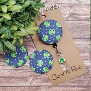 Puede incluir: Tres botones de tela azul y verde con tem&aacute;tica de rana con corazones rosas. Los botones est&aacute;n unidos a una tarjeta marr&oacute;n con el texto "Scout & Remy".