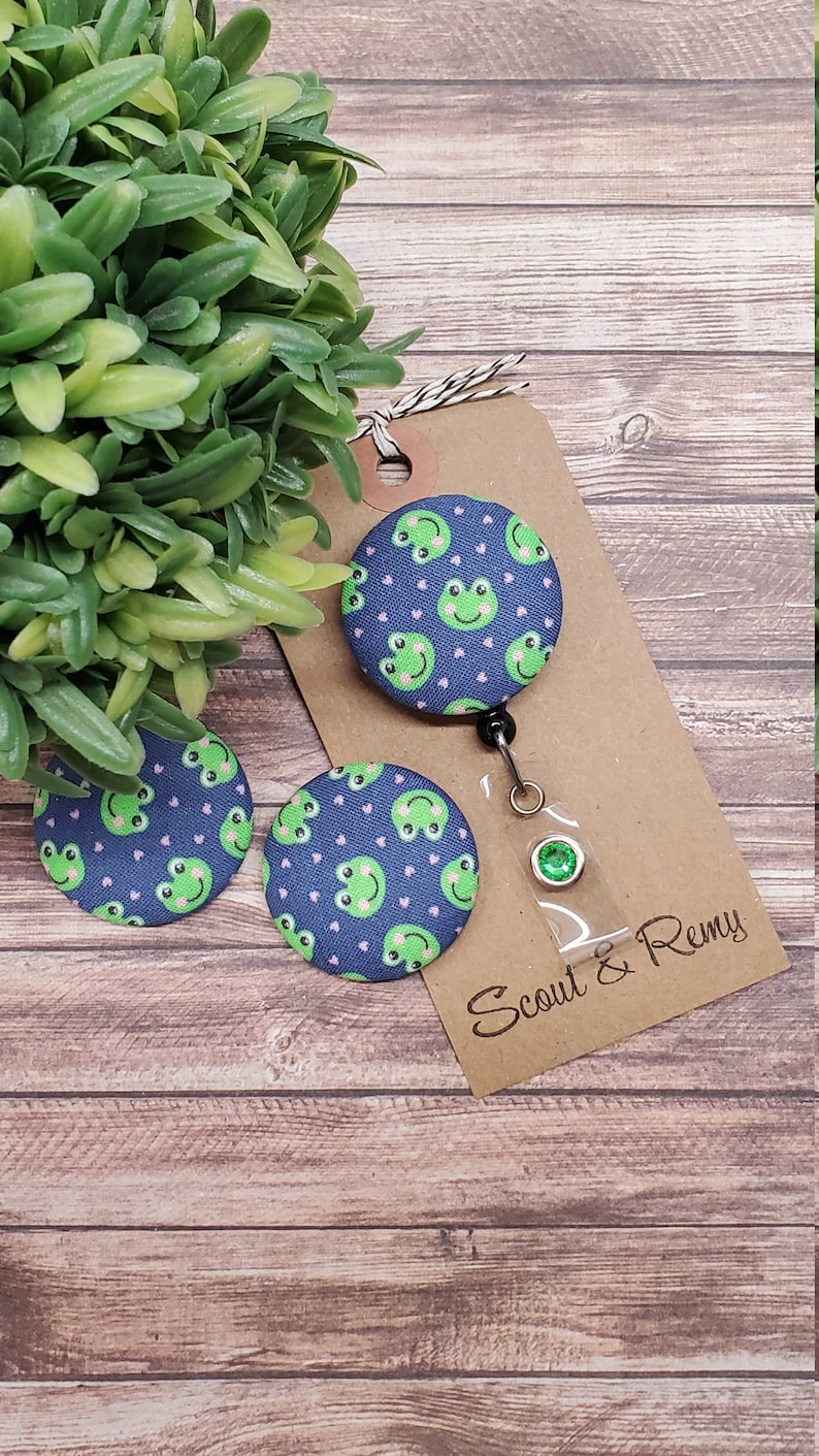 Puede incluir: Tres botones de tela azul y verde con tem&aacute;tica de rana con corazones rosas. Los botones est&aacute;n unidos a una tarjeta marr&oacute;n con el texto "Scout & Remy".