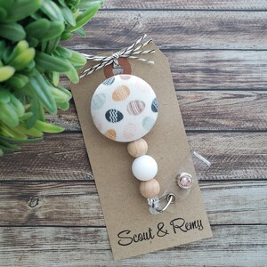 May include: A white and brown beaded badge reel with a patterned fabric button featuring a white background with colorful polka dots. The badge reel is attached to a clear plastic badge holder with a pink rhinestone. The tag below the badge reel reads "Scout & Remy".