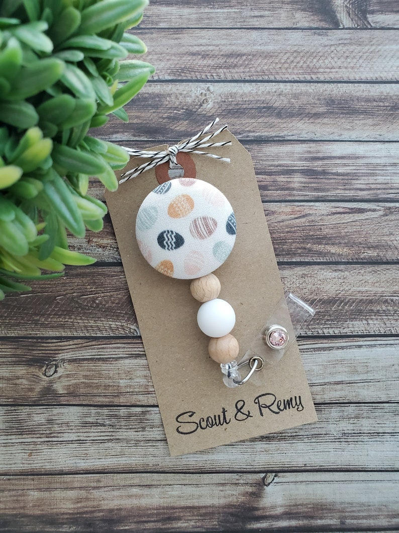 May include: A white and brown beaded badge reel with a patterned fabric button featuring a white background with colorful polka dots. The badge reel is attached to a clear plastic badge holder with a pink rhinestone. The tag below the badge reel reads "Scout & Remy".