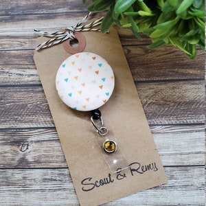 May include: A white fabric badge reel with a colorful heart pattern. The reel has a black retractable cord and a clear plastic badge holder with a gold-colored button. The reel is attached to a brown card with the text "Scout & Remy" written in black.